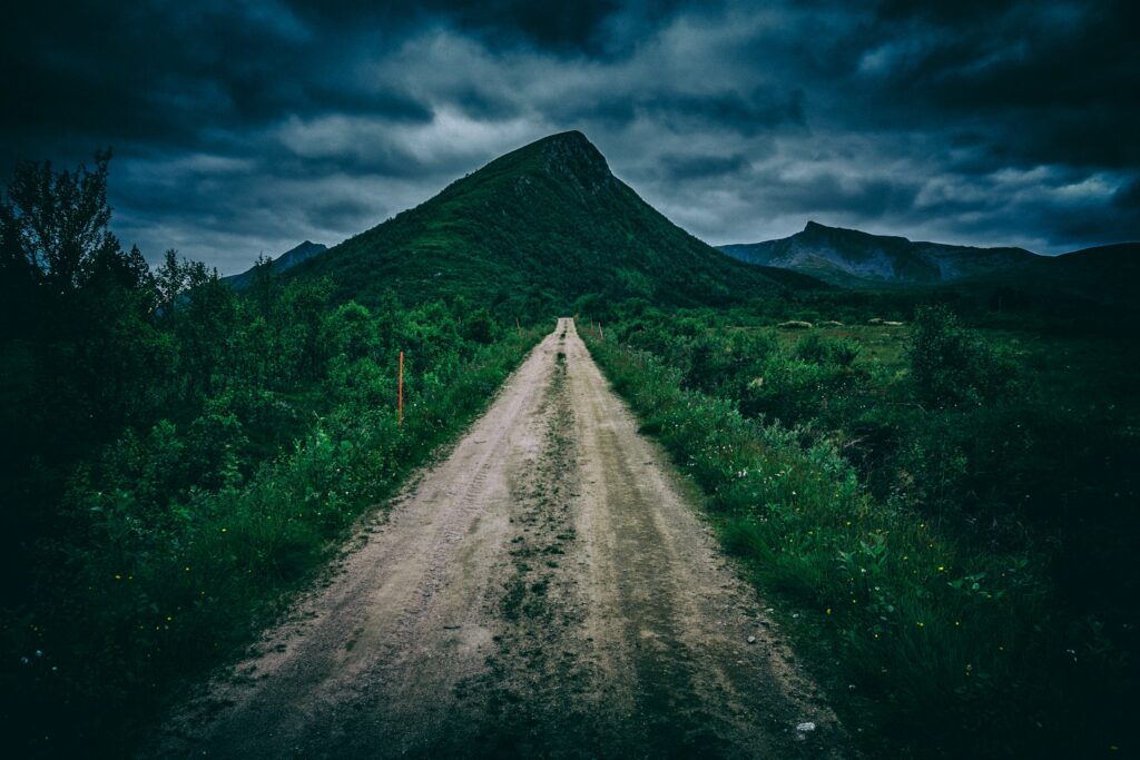 Dramatic dirt path through lush green grass toward striking mountain under moody cloudy sky Wallpaper
