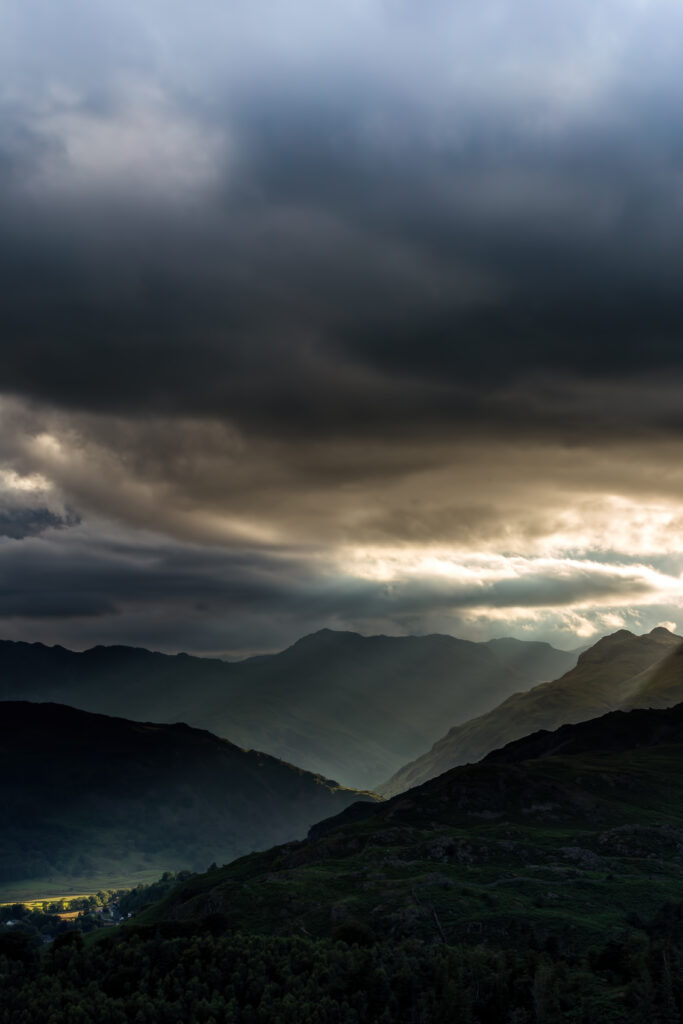 Dramatic mountain landscape with dense forests under moody sky and sun rays breaking through clouds Wallpaper