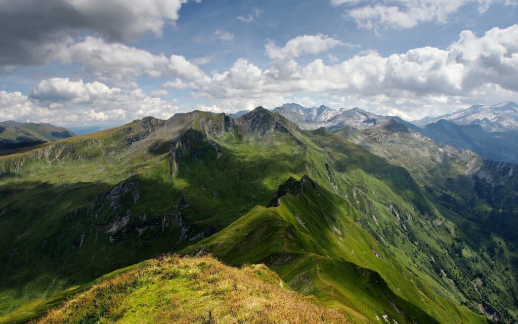 Dramatic mountain landscape with rugged peaks lush greenery deep shadows and vivid blue sky clouds Wallpaper