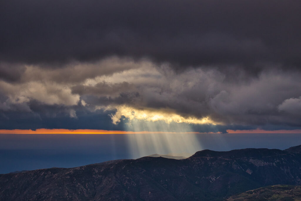 Dramatic mountain landscape with sun rays breaking dark clouds illuminating rugged terrain wallpaper