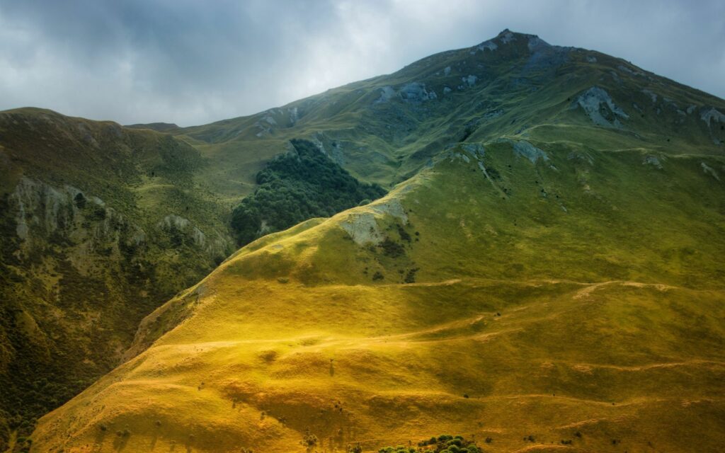 Dramatic mountain landscape with sunlit grassy slopes rugged peaks and moody sky wallpaper background