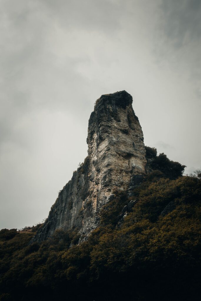 Dramatic towering rock peak above lush forest under moody overcast sky natural landscape wallpaper