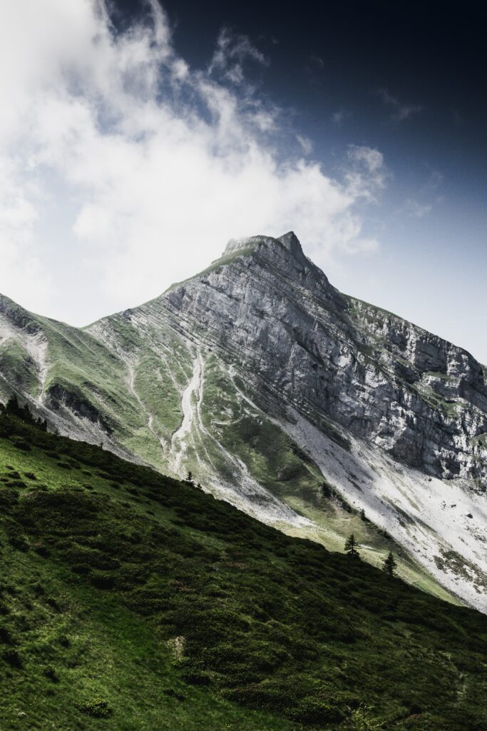 Breathtaking mountain peak above green slopes and dark ridges under swirling clouds and blue sky Wallpaper