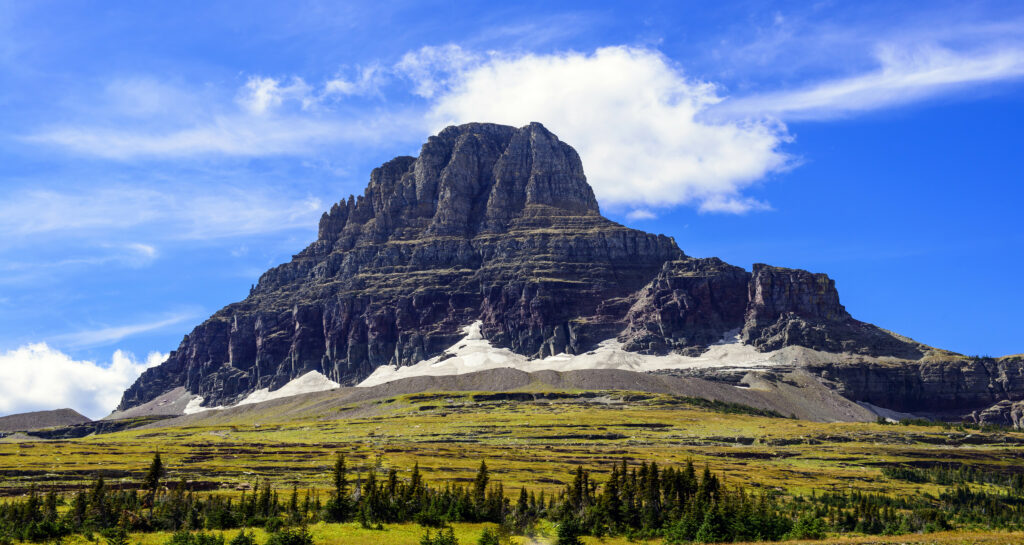 Dramatic rocky mountain peak above green meadows and scattered trees under blue sky clouds Wallpaper