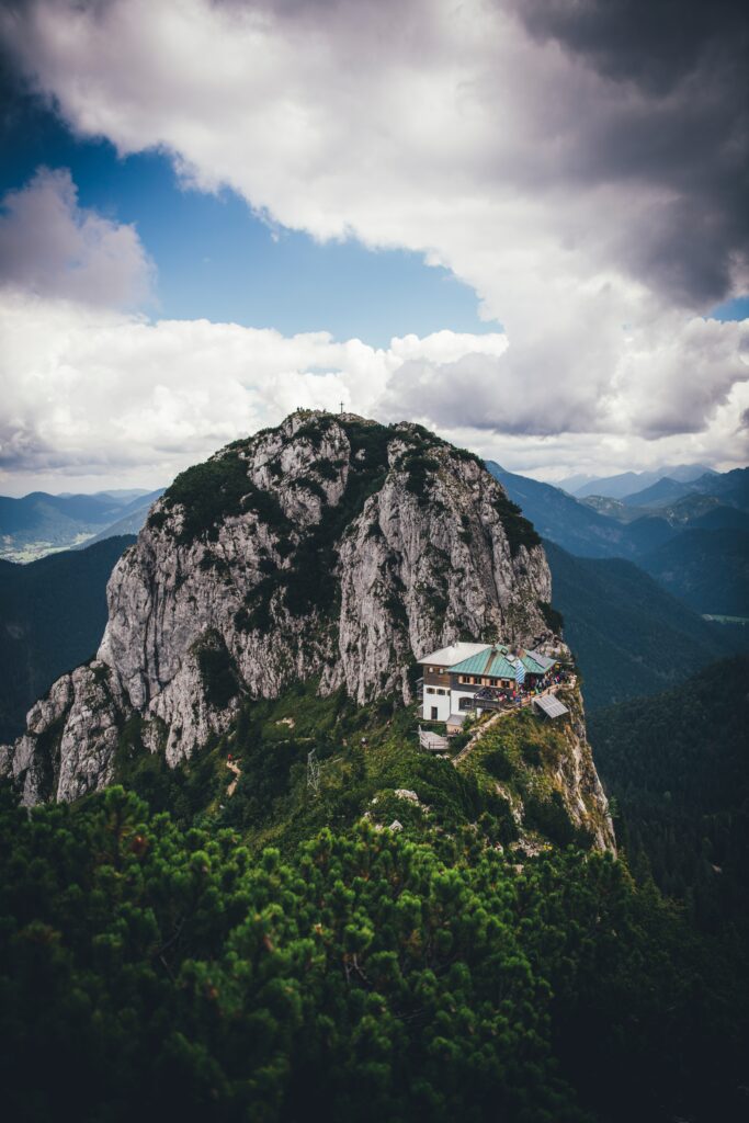 Dramatic mountain peak over green forest with quaint building under dynamic cloudy sky wallpaper