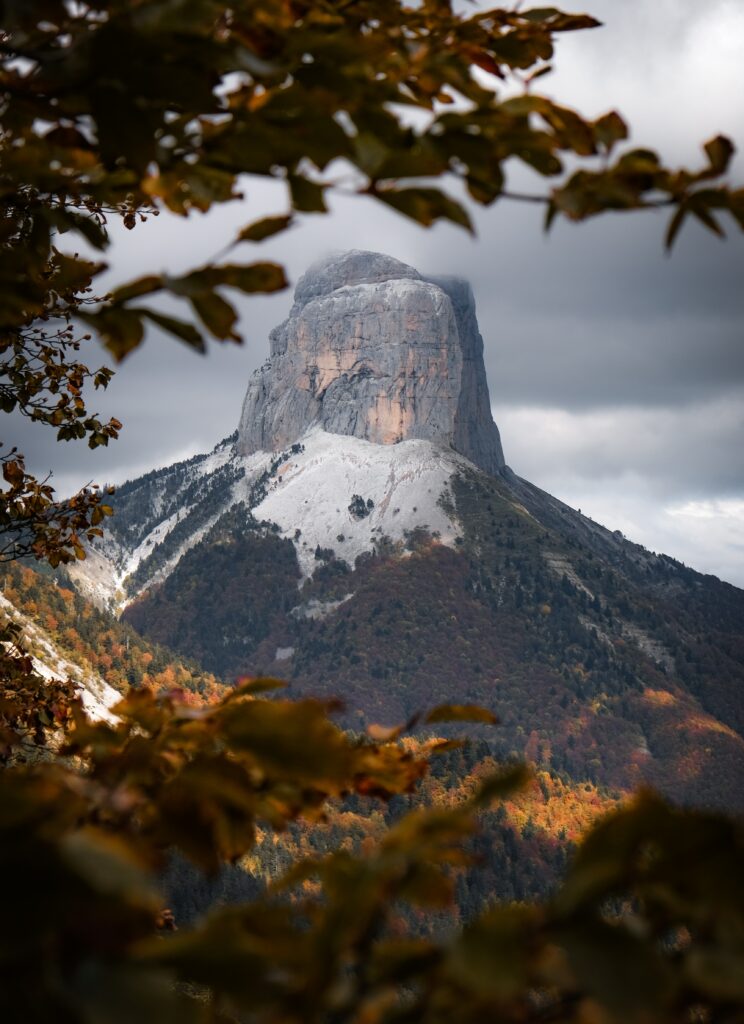 Dramatic mountain peak with snowy cliffs and colorful autumn trees under moody cloudy sky wallpaper