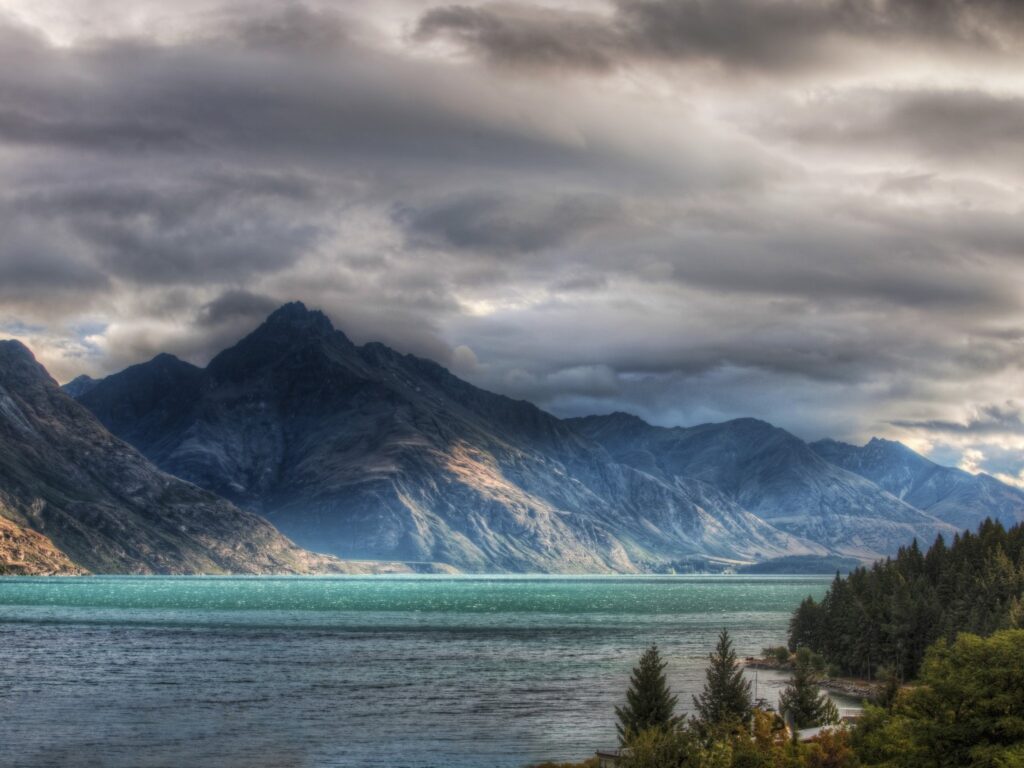 Dramatic mountain peaks over colorful lake with evergreen trees and textured rocks wallpaper background