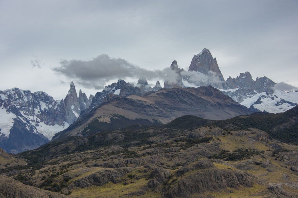 Dramatic mountain range with sharp peaks cloudy sky rugged alpine terrain green brown gray tones Wallpaper