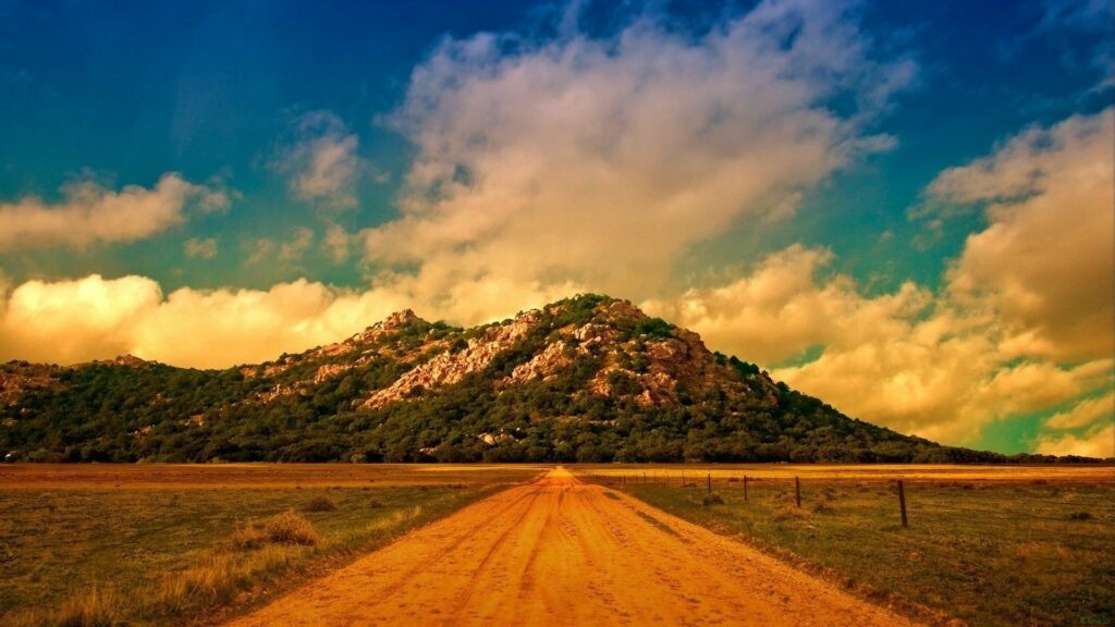 Straight dirt road to rocky mountain through grassy fields under dramatic vibrant sky wallpaper