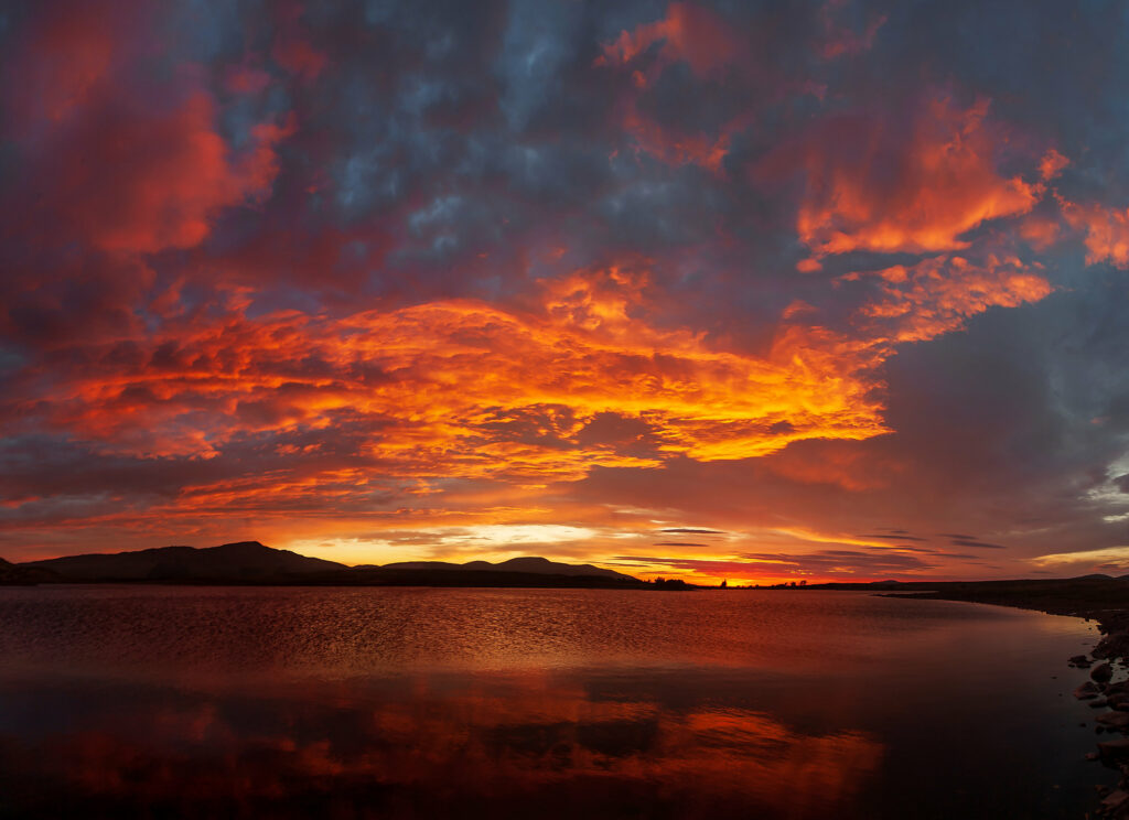 Dramatic mountain silhouette at sunset with fiery orange and blue clouds reflected on calm sea Wallpaper