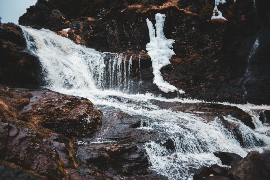 Dramatic mountain waterfall flowing over dark stones with ice patches and stormy sky background Wallpaper