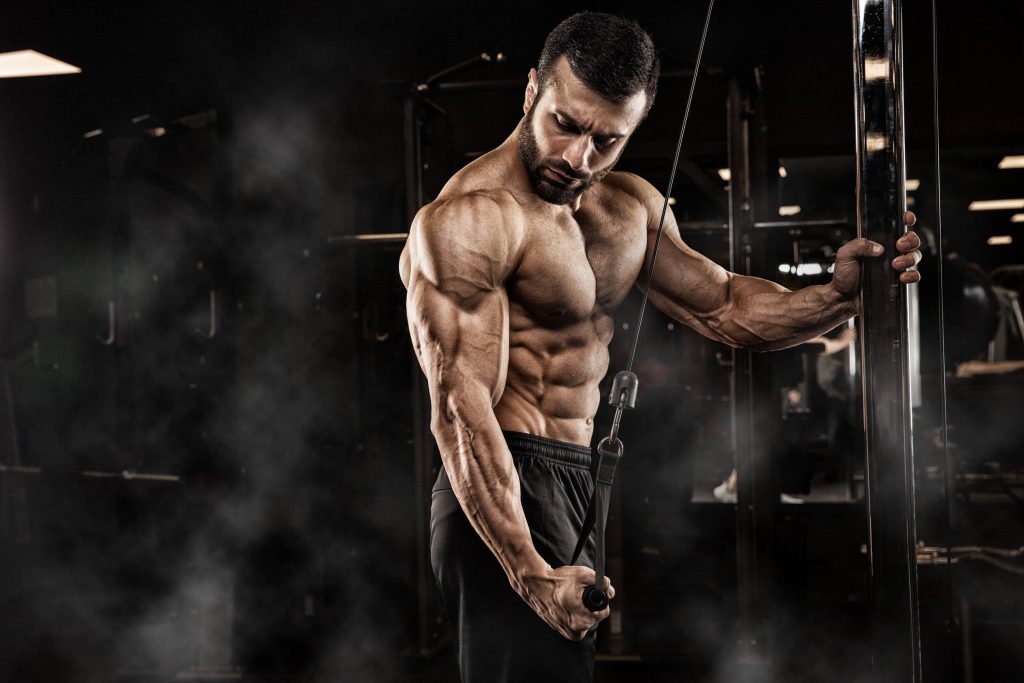 Muscular bearded athlete doing single-arm tricep pushdown in smoky gym with intense lighting Wallpaper