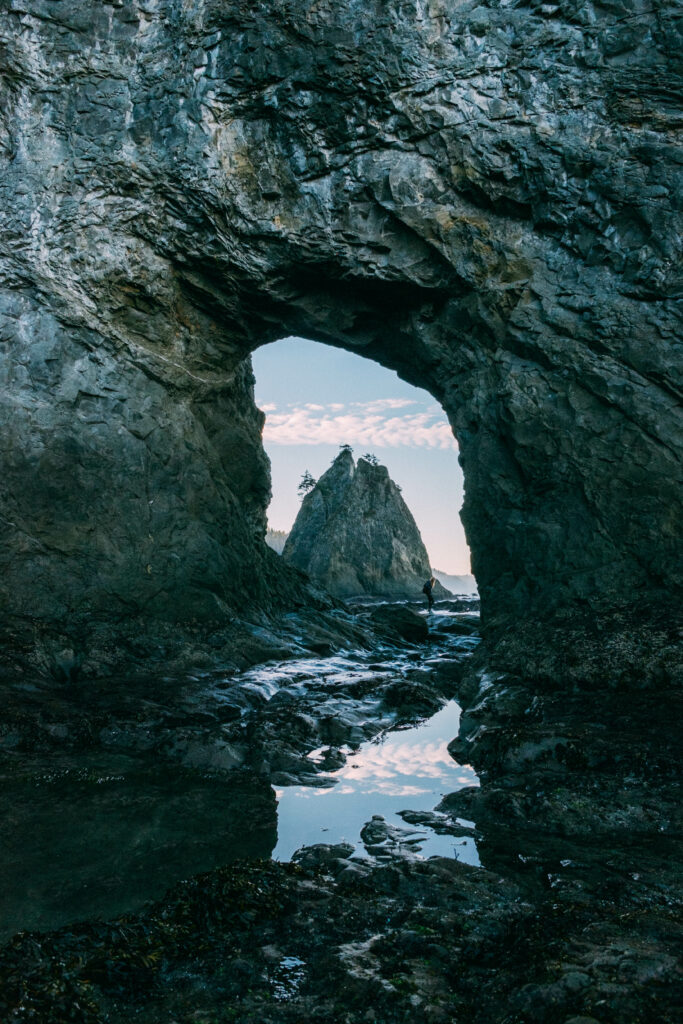 Rugged rock arch framing pointed rock and water reflection with lone silhouette in nature wallpaper