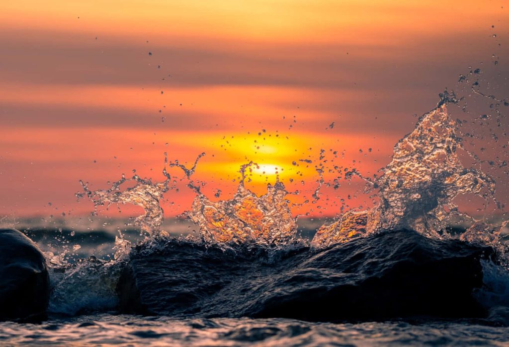 Dramatic ocean splash on dark coastal rocks illuminated by golden sunset sky wallpaper background