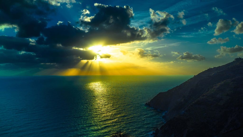 Dramatic ocean sunset with golden rays over deep blue sea and rugged coastline 4K wallpaper