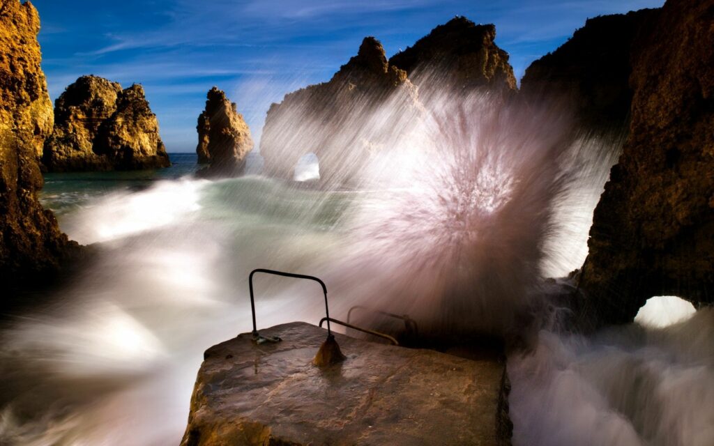 Ocean wave crashing on jagged coastal rocks with water splashes mist under blue sky wallpaper