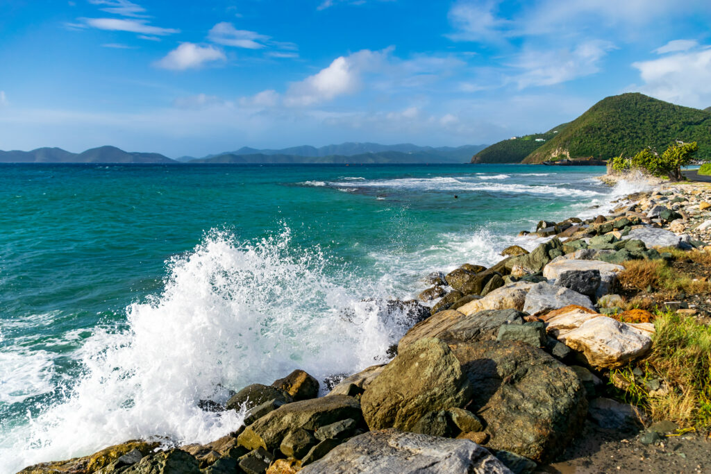 Vibrant ocean wave crashing on rugged stones with splash under blue sky and green hills wallpaper