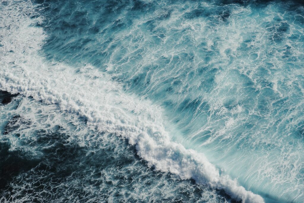 Aerial view of powerful ocean waves crashing with swirling foam in vibrant blues and turquoise Wallpaper