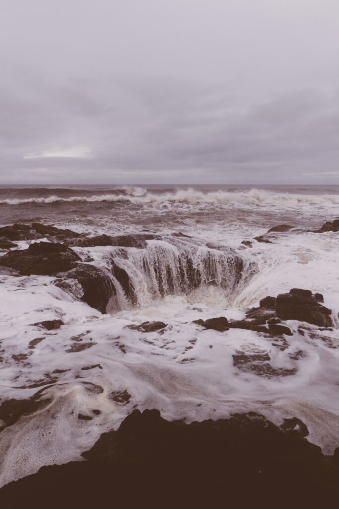 Dramatic ocean waves crash over dark rocks with swirling foam and moody grey brown sky background Wallpaper