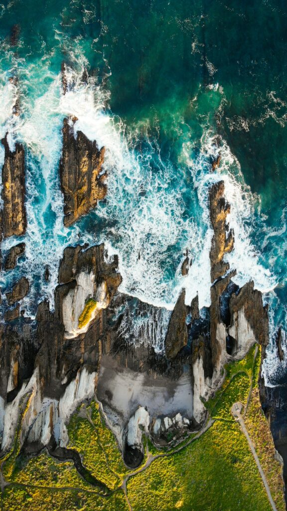Aerial view of crashing ocean waves on rocky coastline with lush greenery vibrant wallpaper background