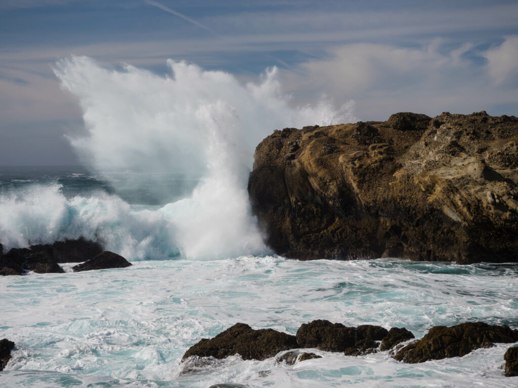 Powerful ocean waves crash against rugged coastal rocks with white splashes under cloudy sky Wallpaper