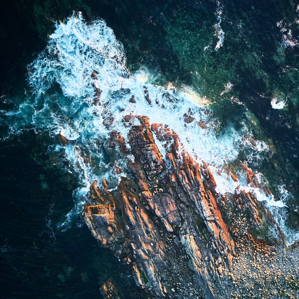 Aerial view of dramatic ocean waves crashing rugged rocks with white sea foam and sunlit stones Wallpaper