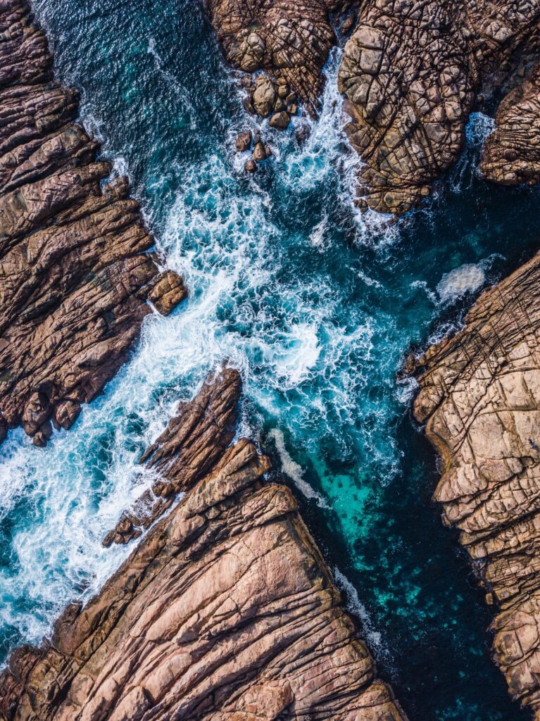 Aerial view of deep blue ocean waves crashing on rugged brown rocks with white splashes wallpaper