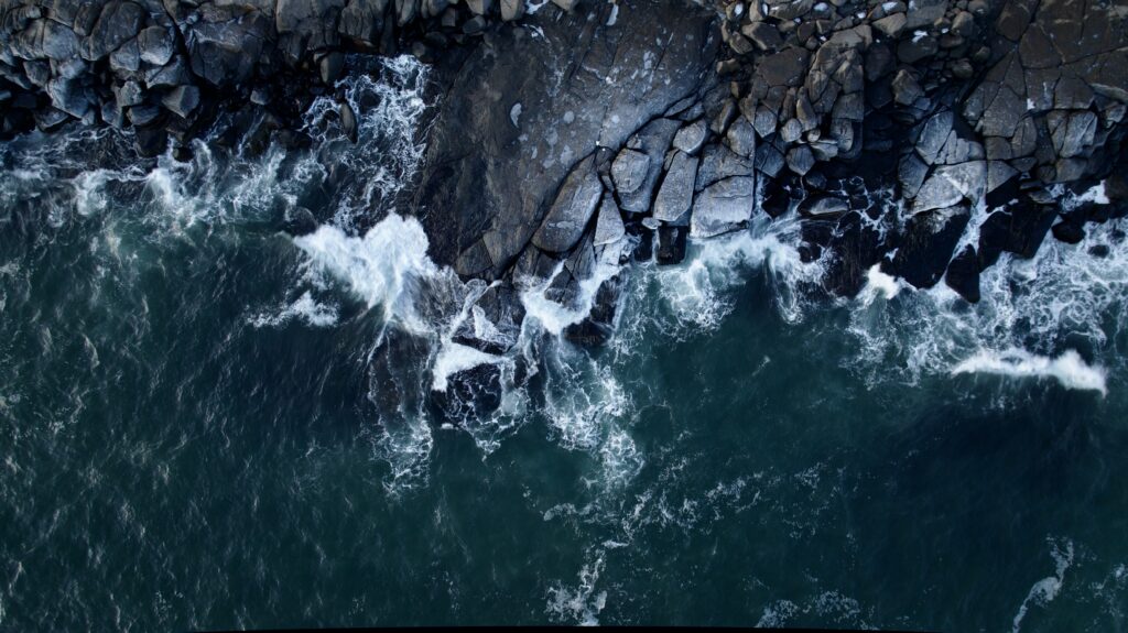 Aerial view of ocean waves crashing on rugged dark rocks with deep blue and turquoise tones wallpaper