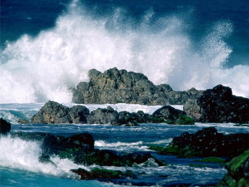 Powerful ocean waves crash against rugged rocks with white splashes and green moss accents Wallpaper