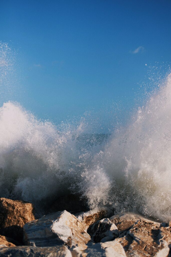 Waves crashing on rugged shore rocks with white surf under clear blue sky natural seascape Wallpaper