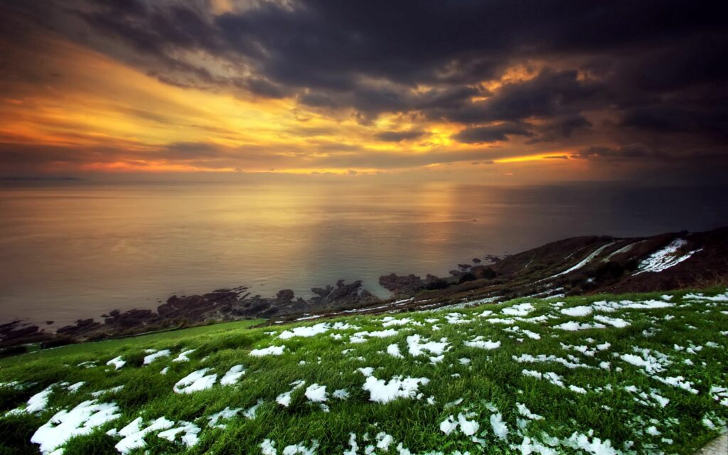 Dramatic orange and gold sunset over calm sea with snowy patches on green coastal hillside wallpaper