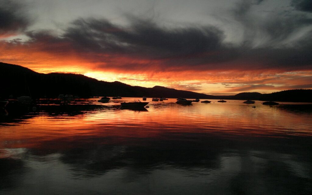 Dramatic sunset wallpaper with orange gray clouds and silhouetted boats reflecting on calm water