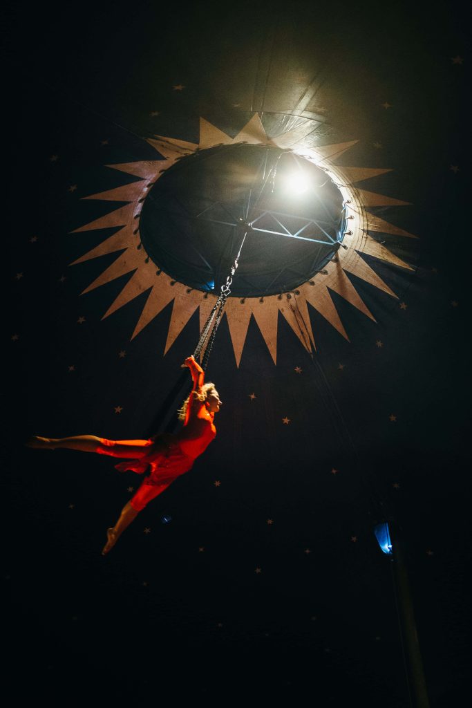 Aerialist in red costume suspended under starry circus tent with golden sun design wallpaper background