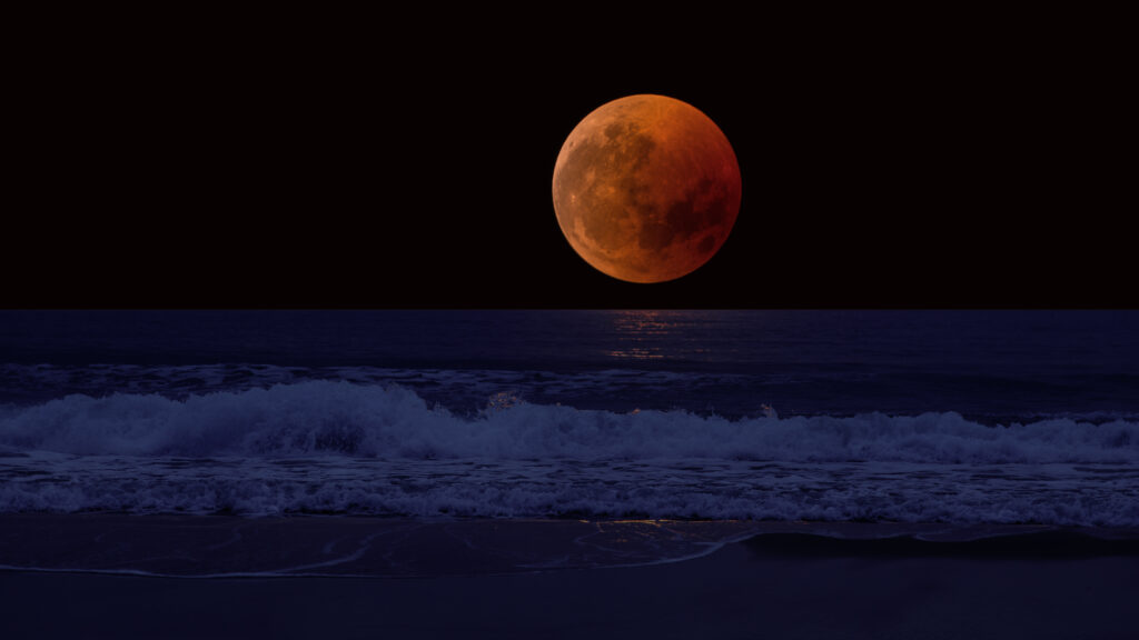 Dramatic red lunar eclipse over dark sea waves gently crashing on serene mystical coast wallpaper background