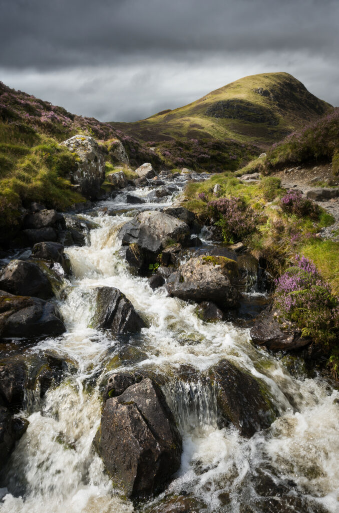 Vibrant river flowing over dark stones through wildflowers and green hills under cloudy skies wallpaper