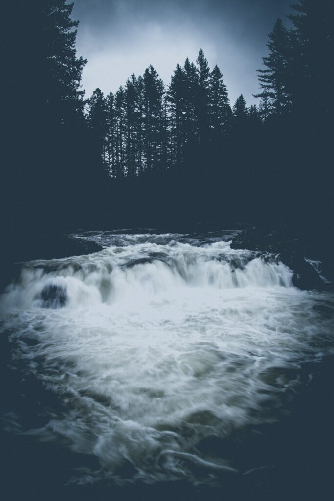 Dramatic river rushing over rocks with tall trees under dark moody sky deep blue tones wallpaper