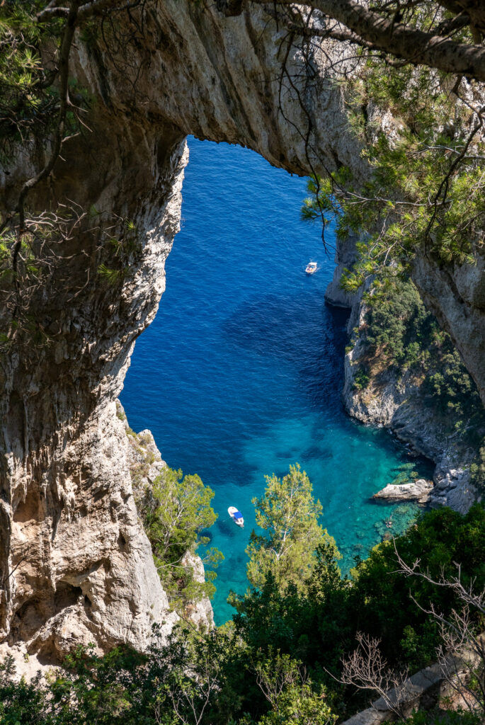 Dramatic rock arch over vibrant blue sea with lush trees and small boats in tranquil coastal scene Wallpaper