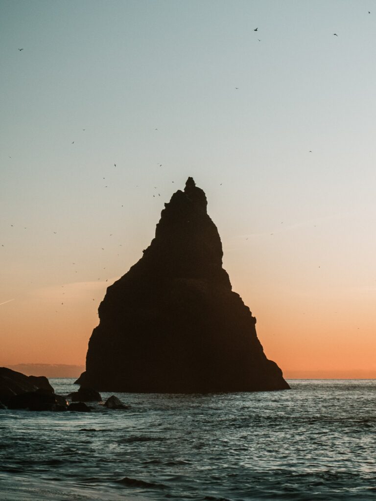 Dramatic rock formation silhouette at dusk over calm sea with orange blue sky and gentle waves Wallpaper