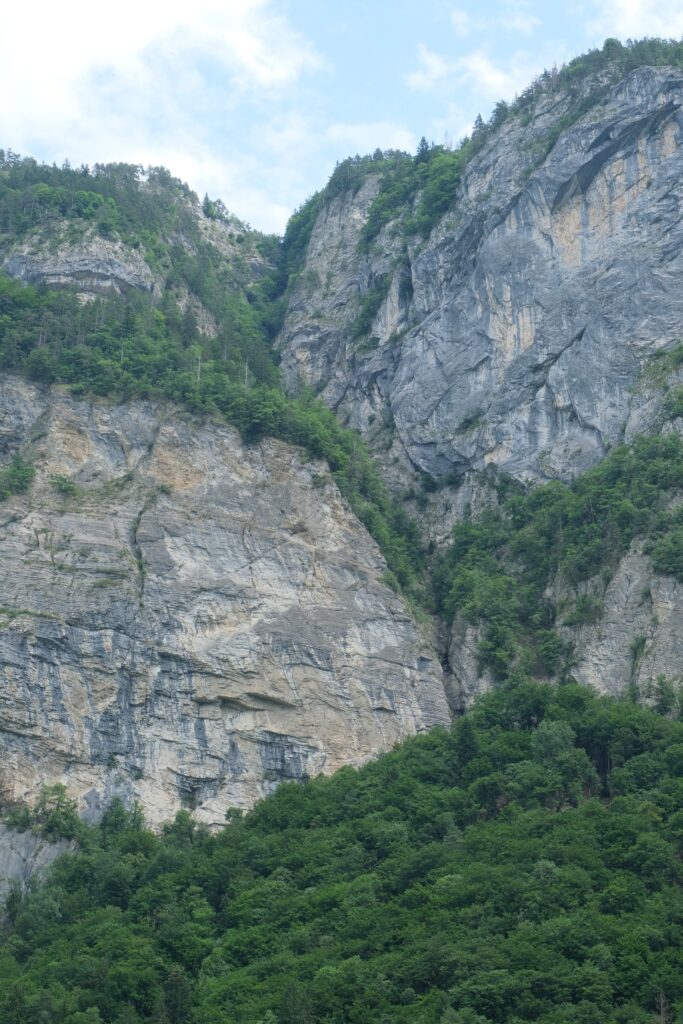 Dramatic rocky cliffs rise above dense green trees with textured stone and pale blue sky wallpaper