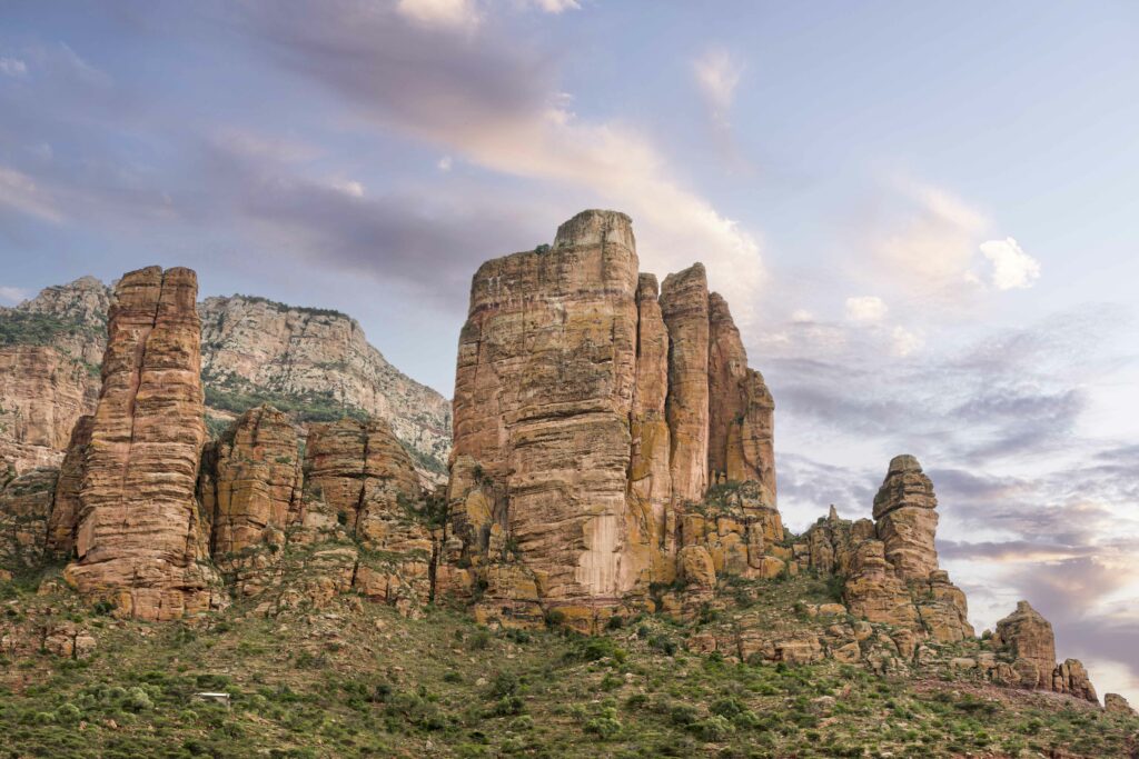 Towering rock formations on rocky slope with green bushes under pastel sky and soft clouds Wallpaper