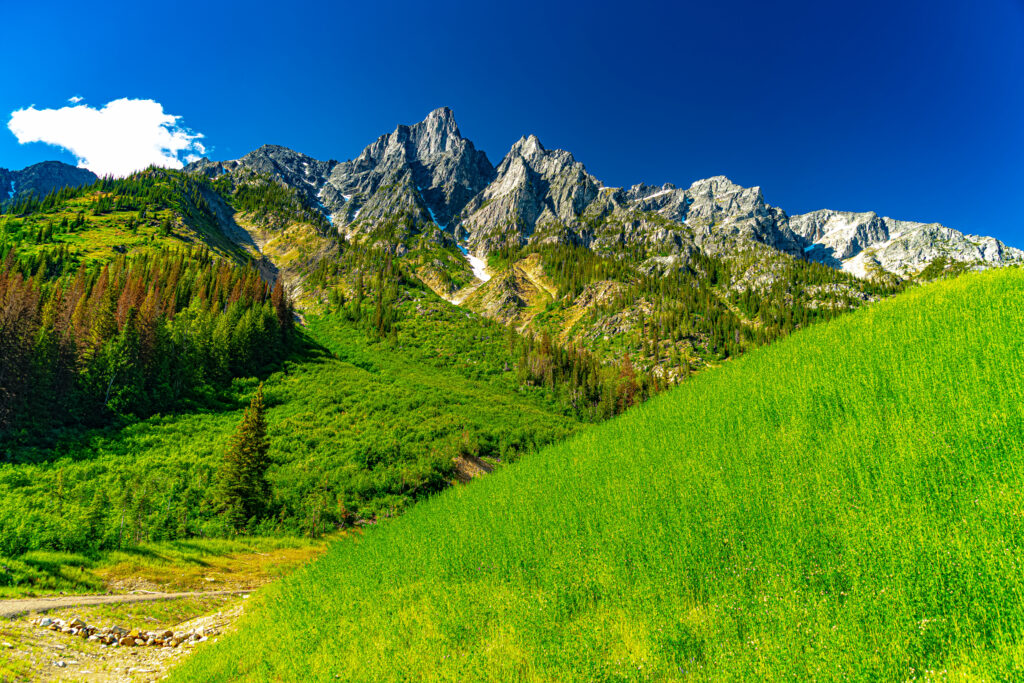 Dramatic rocky mountains above lush green valley with vibrant grass fields under blue sky wallpaper