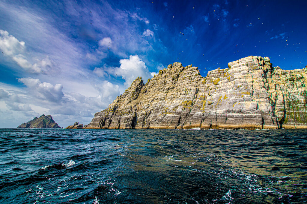 Rugged cliffs above crashing sea waves under vibrant blue sky with scattered clouds wallpaper