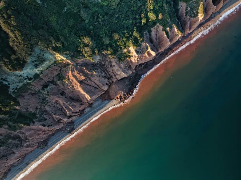 Aerial view of rugged rocky coastline meeting serene sea with green and reddish-brown tones wallpaper