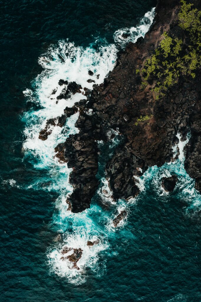 Aerial view of rugged coastline with dark rocks white surf and vibrant blue sea and cliffs greenery Wallpaper