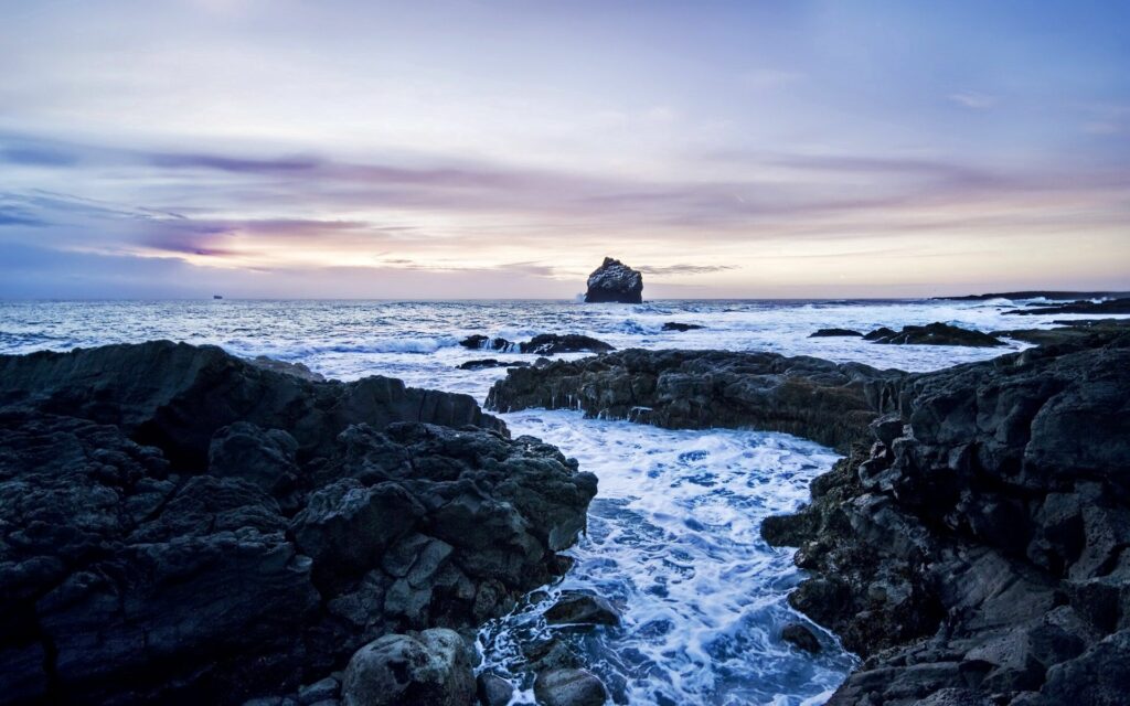 Rugged coastline with dark rocks foamy waves under cloudy evening sky dramatic cold seascape Wallpaper