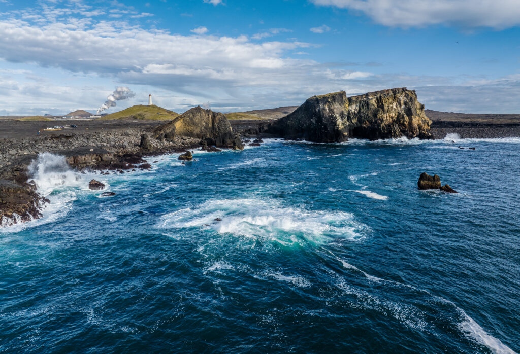 Rugged coastline with crashing waves lighthouse on grassy hill under vibrant blue sky wallpaper
