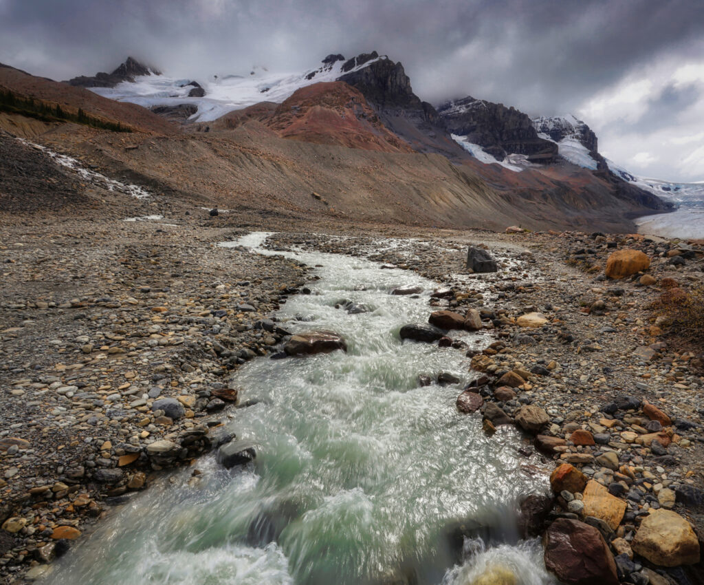 Rugged snow-capped mountains with winding stream under moody sky wilderness landscape wallpaper