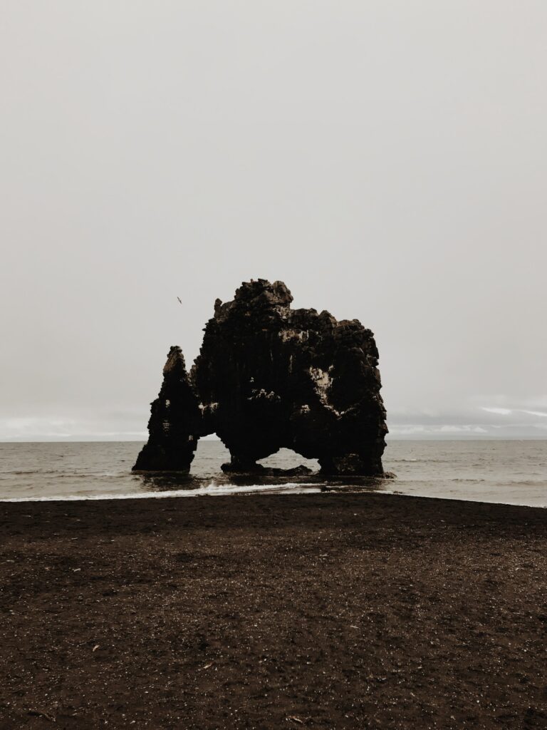 Dramatic rugged rock formation on dark sandy beach under cloudy sky tranquil seascape wallpaper