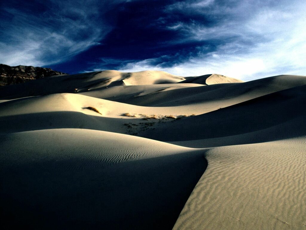 Sweeping desert sand dunes with shadows under blue sky wispy clouds and distant mountains background Wallpaper