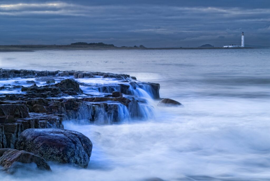 Dramatic Scottish coast with rugged stones and crashing waves near solitary beacon under cloudy sky Wallpaper